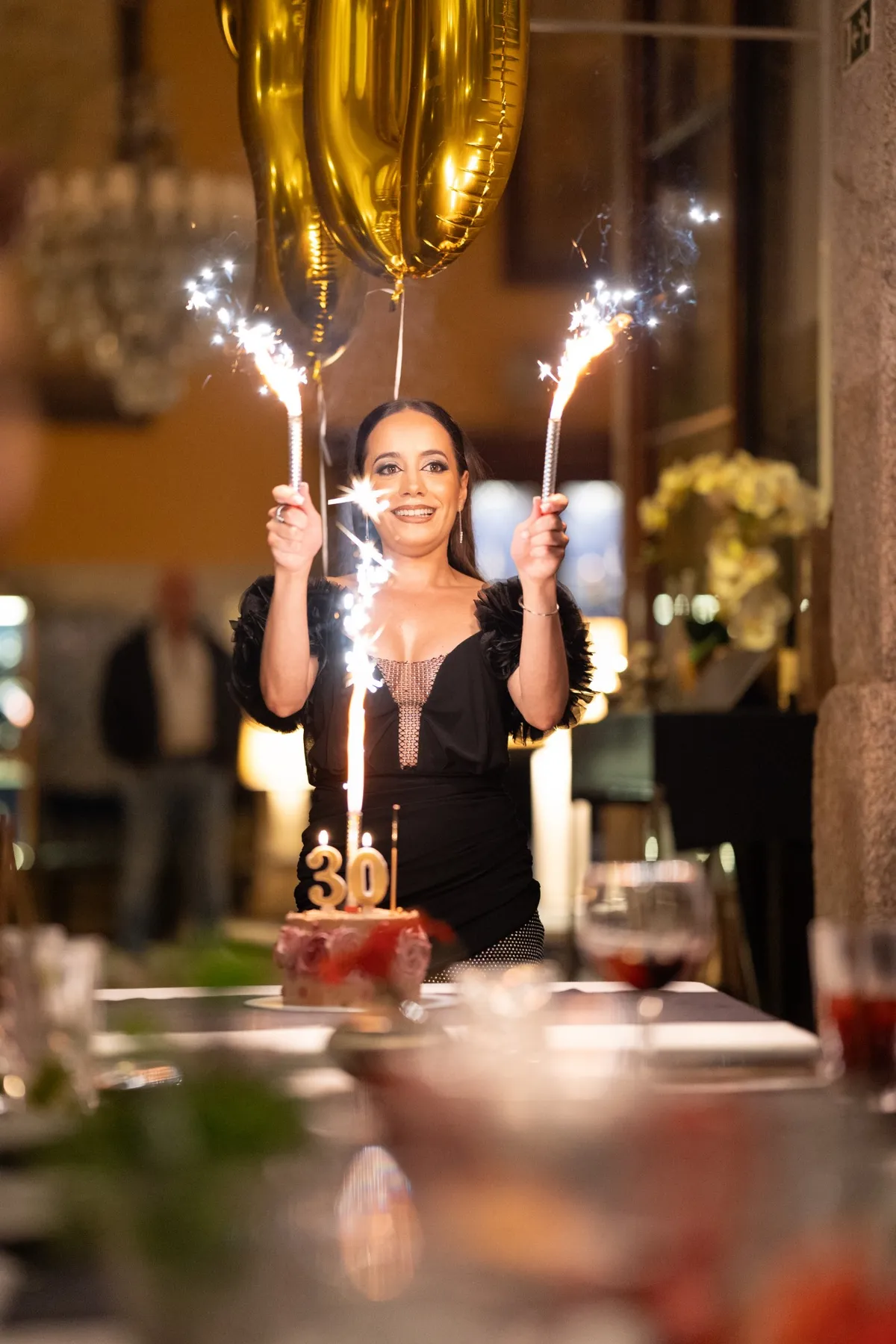 Mulher de vestido preto a sorrir, com dois fogos de artifício de mesa e balões dourados com o número 30