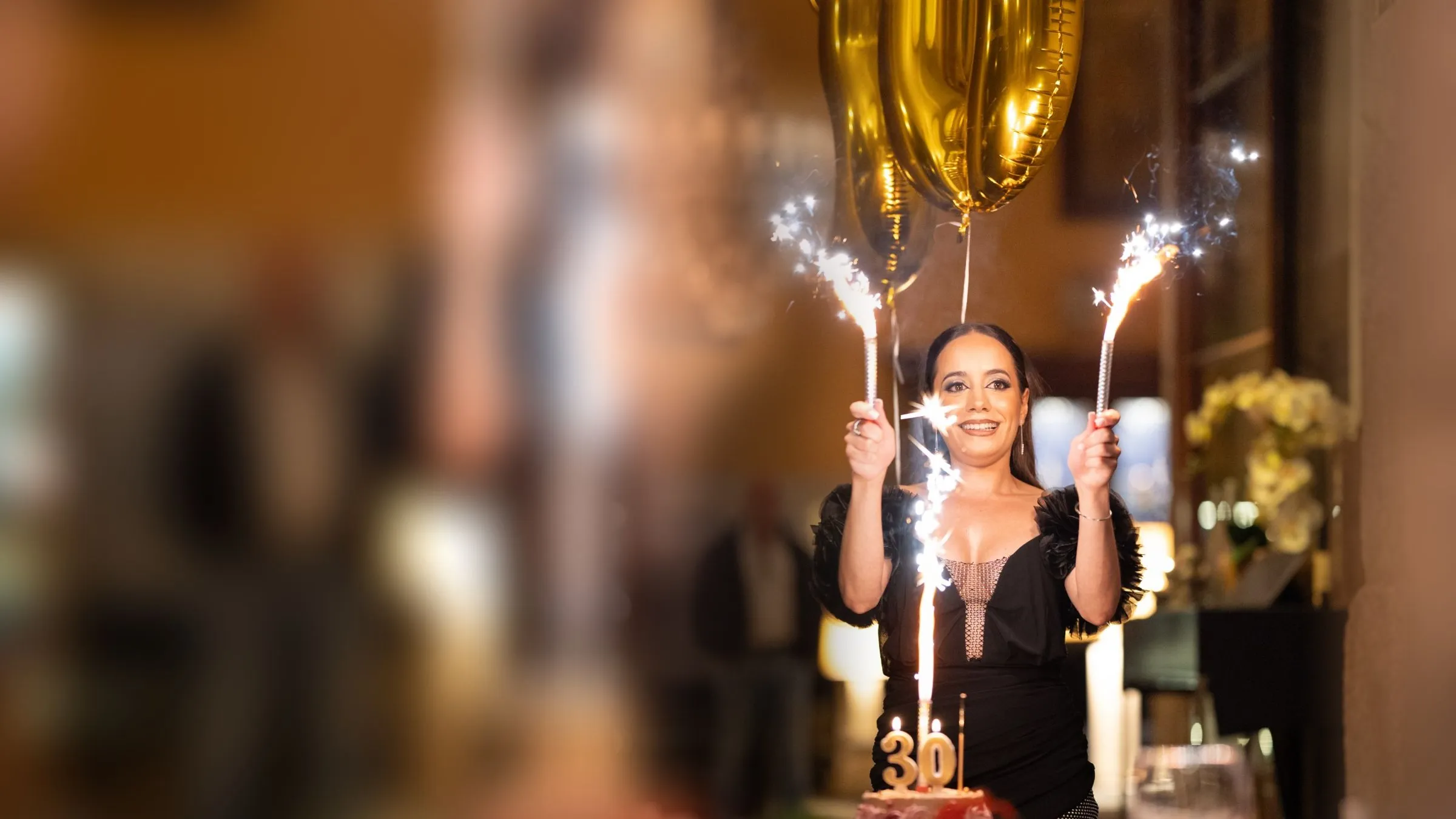 Mulher de vestido preto a sorrir, com dois fogos de artifício de mesa e balões dourados com o número 30