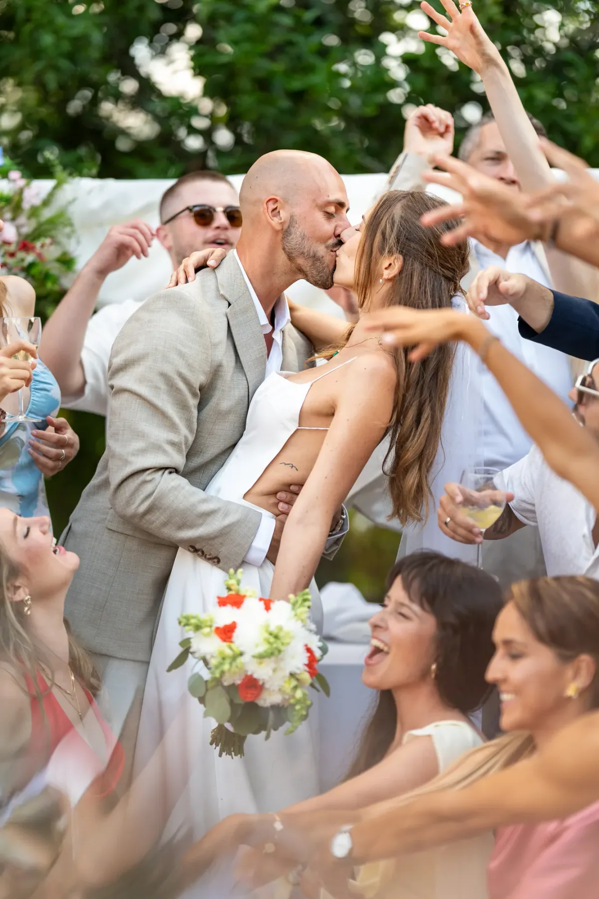 Noivos a beijar-se rodeados de convidados a festejar, com o ramo de noiva em primeiro plano