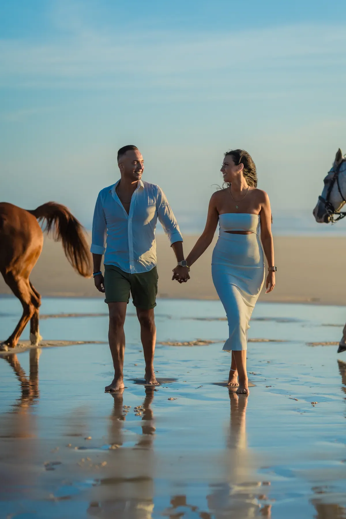 Casal a caminhar de mãos dadas na praia ao entardecer, com dois cavalos