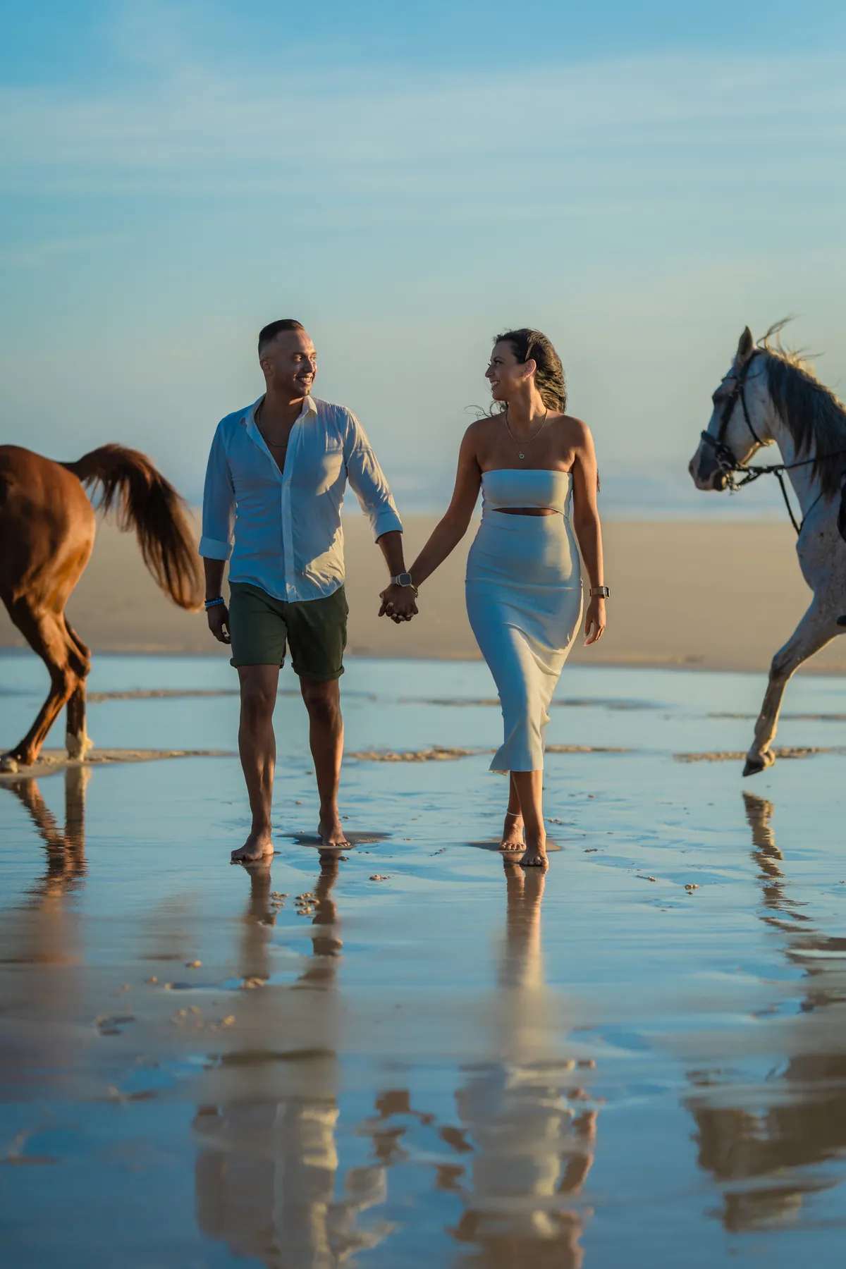 Casal a caminhar de mãos dadas na praia ao entardecer, com dois cavalos