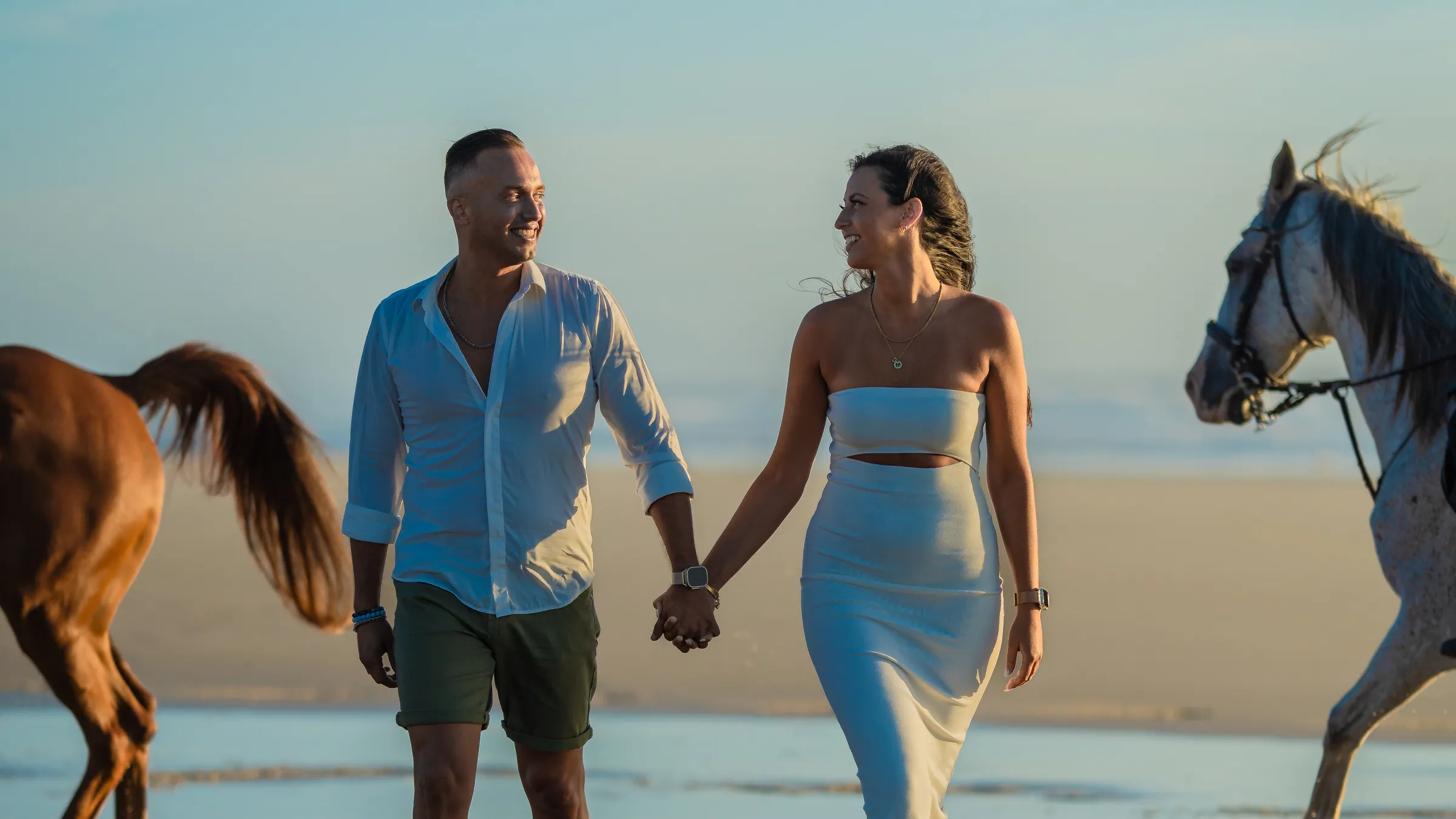 Casal a caminhar de mãos dadas na praia ao entardecer, com dois cavalos